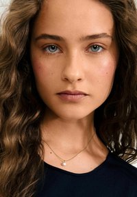Collier en chaîne dorée avec un petit pendentif en perle. Le mannequin a des cheveux ondulés, brun moyen et porte un haut sombre avec un col rond.