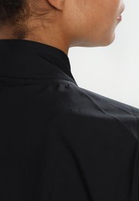 Chemise noire avec un col montant, fabriquée en tissu lisse. Présente un design épuré et minimal mis en valeur par des détails de couture subtils.