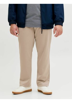 Uomo con pantaloni beige, scarpe da ginnastica bianche, maglietta grigia e giacca blu scuro in piedi su sfondo bianco.