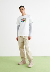 Camiseta blanca con un estampado gráfico colorido, superpuesta a una camiseta de manga larga gris claro. Pantalones cargo beige con puños elásticos y detalles mínimos.