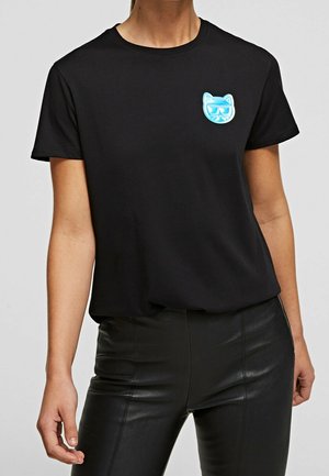 Černé tričko s krátkým rukávem a malou modrou nášivkou kočičí hlavy na levé straně hrudi, nošené s černými koženými kalhotami.