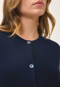 Marineblauwe gebreide cardigan met een ronde halslijn en een enkele lichtblauwe knop in het midden. Zachte textuur en eenvoudig ontwerp.