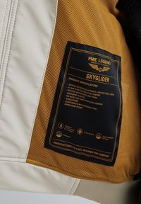 Softshell jacket featuring a beige exterior, mesh lining in gold, and a black label detailing fabric composition and product specifications.