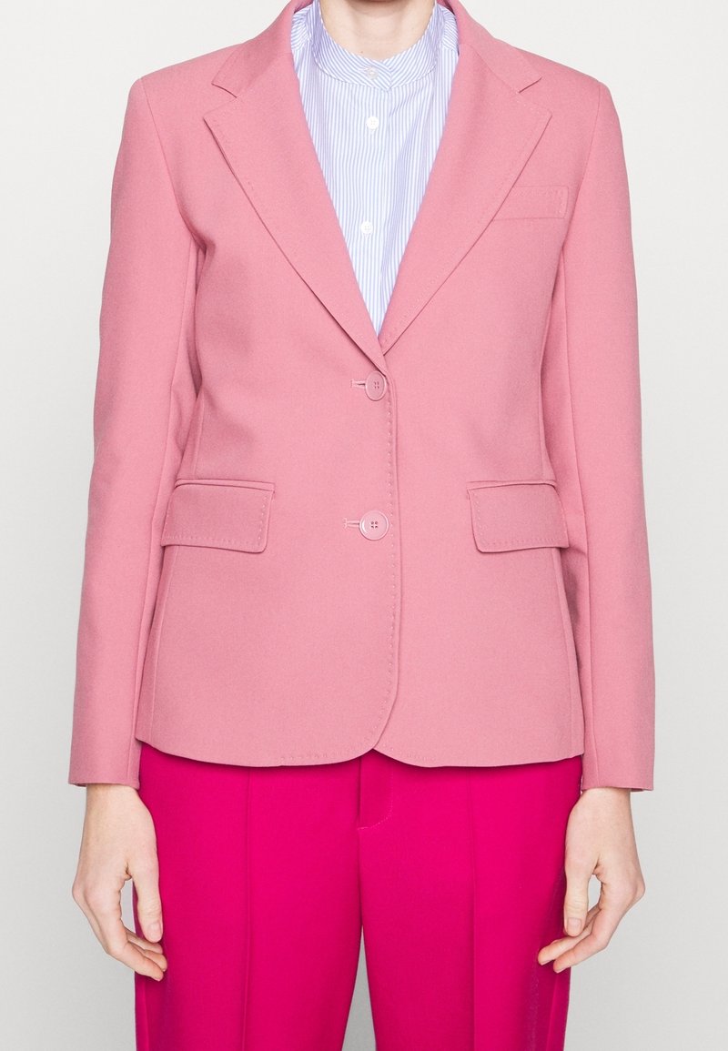 Blazer rosa claro con dos botones y bolsillos laterales, hecho de una tela suave. Llevado sobre una camisa a rayas azul y blanca, con un cuello estrecho.