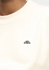 Κοντινό πλάνο ενός ατόμου που φοράει λευκό μπλουζάκι με στρογγυλή λαιμόκοψη και μικρό, σκοτεινό ημικυκλικό λογότυπο στο αριστερό στήθος.