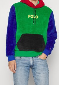 Měkká, vícebarevná mikina s kapucí se zelenými, modrými a červenými panely, uprostřed černá kapsa a vyšívané logo "POLO" žluté barvy.