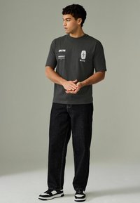 Un tricou gri din bumbac, cu mâneci scurte, prezentând imprimeuri grafice albe; purtat cu blugi negri din denim și adidași negri/albi.