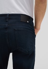 Ciemnoniebieskie dżinsy o dopasowanym kroju. Posiadają czarną skórzaną metkę w talii oraz tylnią kieszeń z zaokrąglonym szwem.