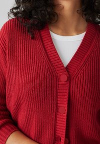 Punainen neulecardigan, jossa on V-pääntie, ribattua pintaa ja kolme pyöreää nappia. Värin päällä valkoinen paita, jossa on näkyviä ompelutietoja.