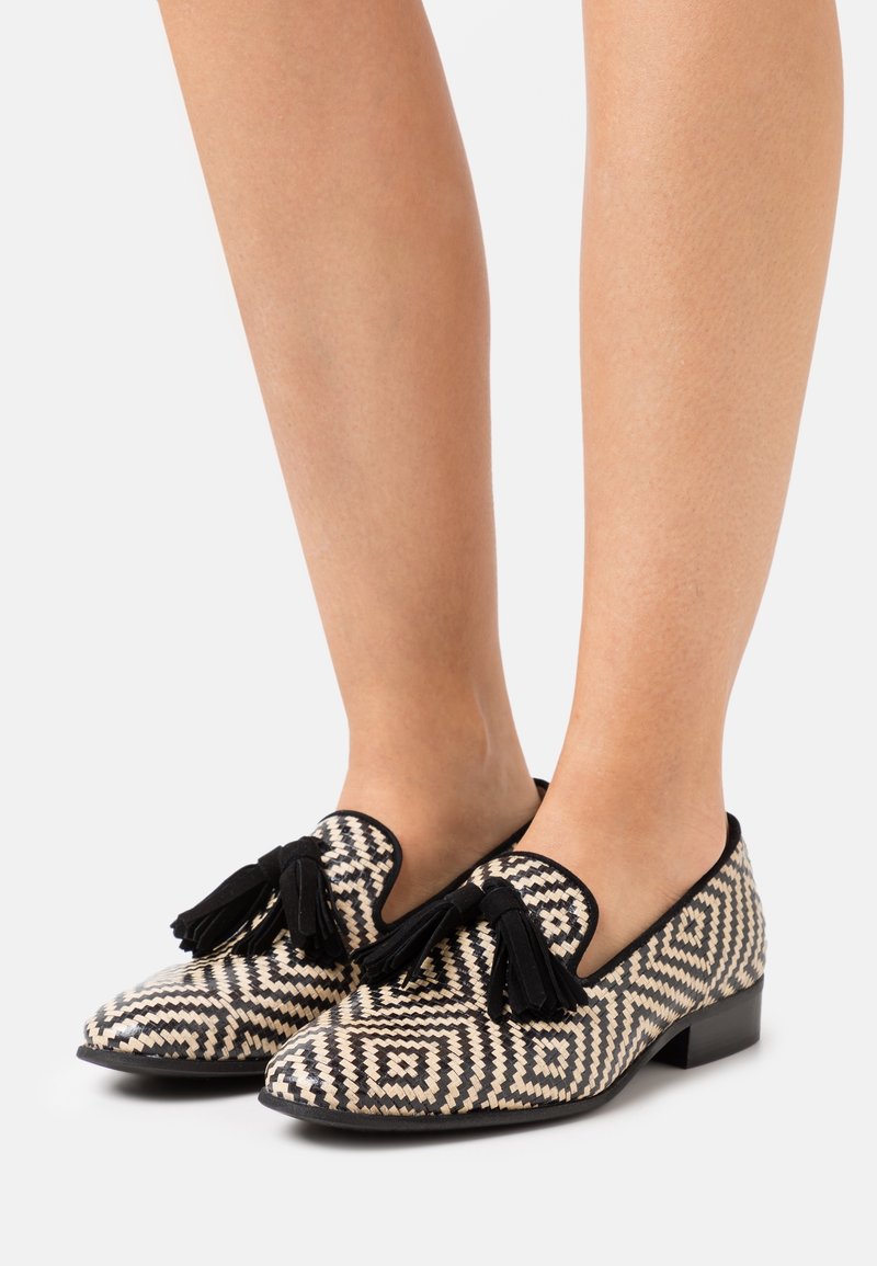 Pieds portant des mocassins à motif géométrique noir et blanc avec des pampilles noires, montrés sur un fond clair uni.