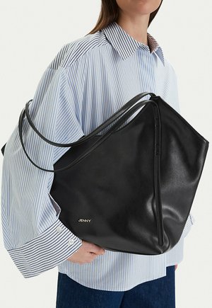 Mujer con una camisa de rayas azules y blancas sosteniendo un gran bolso tote de cuero negro con tirantes delgados para el hombro y el logo "JENNY".