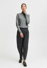 Pull en col roulé gris associé à un pantalon plissé sombre. Le mannequin porte des mocassins noirs avec des accents dorés. Texture de tissu lisse, couleurs neutres.