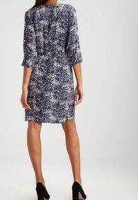 Kleid in Marineblau mit einem weißen abstrakten Muster, mit mittellangen Ärmeln und einer taillierten Passform. Abgerundet mit schwarzen High Heels.