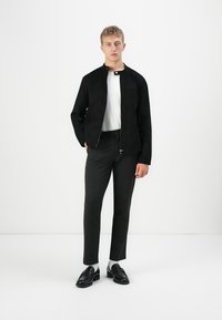 Un jeune homme se tenant debout, portant une veste zippée noire, une chemise blanche, un pantalon slim noir, des chaussettes blanches et des mocassins noirs, devant un fond blanc uni.