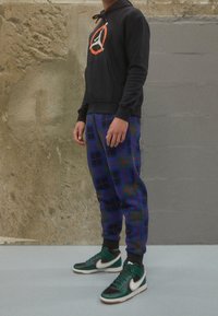 Sudadera negra con un logo, pantalones jogger de cuadros azules con acentos verdes, y zapatillas altas negras/verde con detalles blancos.