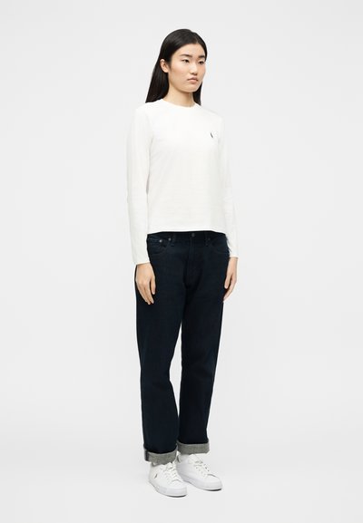 Νεαρή γυναίκα όρθια, φοράει άσπρο πουκάμισο με  μακριά μανίκια, σκουρόχρωμα τζιν με κουμπωμένα μανίκια και άσπρα αθλητικά παπούτσια, πάνω σε απλό φωτεινό φόντο.