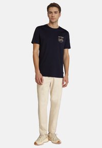 Camiseta de algodón azul marino con mangas cortas, con un logotipo gráfico dorado en el bolsillo del pecho, combinada con pantalones beiges y zapatillas.
