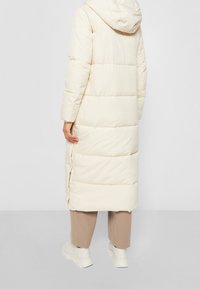 Lang, cremefarvet polstret frakke med hætte. Har vandrette syninger, sidelæg og en glat, shiny tekstur.