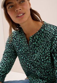 Femme portant un haut à manches longues à motif teal et noir et une casquette de couleur claire, assise contre un fond léger uni.