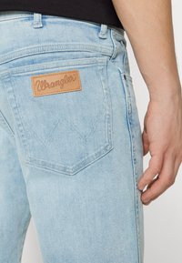 Gaiši zili džinsi ar ādas Wrangler uzlīmi uz aizmugurējā jostas, ar izteiktu 'W' šuvju dizainu uz kabatām.