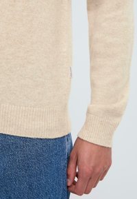 Maglione lavorato a maglia beige con polsini e orlo a coste, texture morbida, vestibilità slim, presenta un piccolo etichetta del marchio sul lato. Abbinato a jeans.