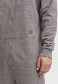 Gray zip-up onesie made of soft, textured fabric. Features a leather patch with "BLEND" logo and ribbed cuffs. Zipper closure at front.
