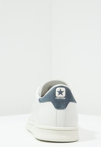 Baskets en cuir blanc avec un talon en daim bleu et un écusson logo, forme arrondie et semelle en caoutchouc texturée. Design épuré et minimaliste.