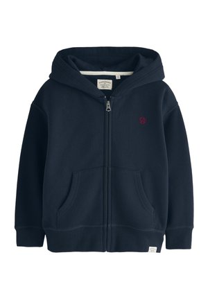 Junger Junge mit hellbraunen Haaren, der einen marineblauen Zip-Hoodie über einem beigefarbenen Shirt trägt, steht vor einer schlichten hellen Wand.