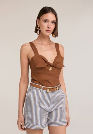 Vrouw met kort bruin haar draagt een bruine mouwloze top met knoop aan de voorkant, gestreepte shorts met hoge taille, een bruine riem en grote oorbellen.