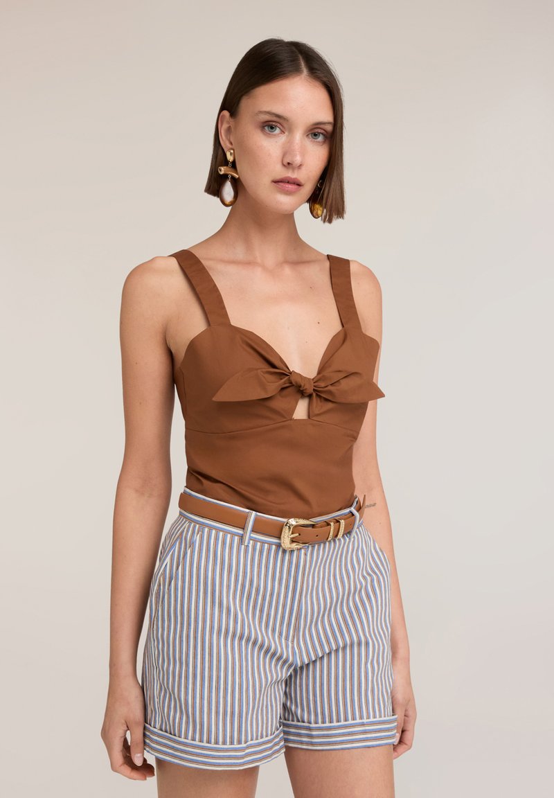 Woman with short brown hair wears brown sleeveless top with front knot, striped high-waist shorts, a brown belt, and large earrings.
