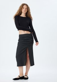 Top noir à manches longues et jupe midi froncée noire à pois avec fente latérale. La jupe présente une texture lisse et une taille plate. Chaussures plates noires.