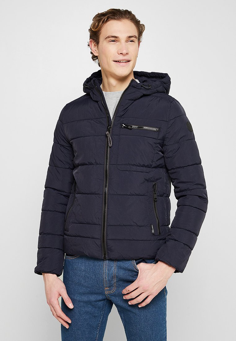 Marineblaue Steppjacke mit durchgehendem Reißverschluss, Kapuze und zwei Seitentaschen. Verfügt über eine gekleidete Textur und eine Brusttasche mit Reißverschlussdetails.