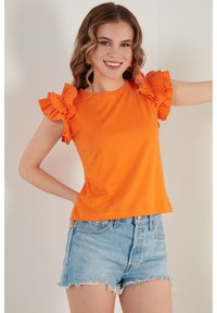 Oranžové bavlněné tričko s volánkovými krátkými rukávy, kombinované se světle modrými džínovými šortkami. Jednoduchý design s kulatým výstřihem.