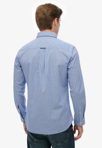 Lyseblå stribet skjorte med knap-ned krave og lange ærmer. Har en central plisse og knapærmer. Stoffet virker glat.