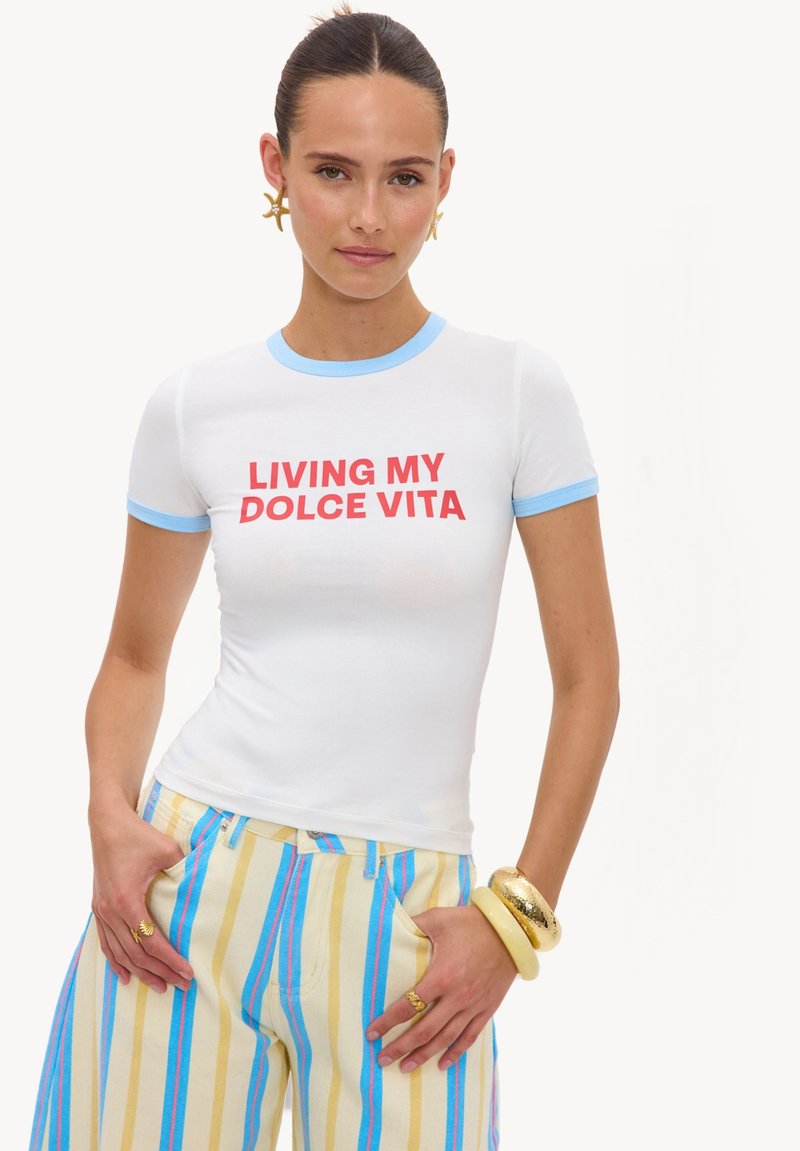Femme aux cheveux foncés portant un t-shirt blanc avec bordure bleue et texte rouge "LIVING MY DOLCE VITA,", portant un pantalon rayé et des bracelets dorés et crème.