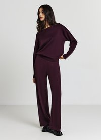 Bordeauxfarbener Strickanzug mit locker sitzendem Pullover und weitem Hosenbein, beide mit einer weichen Textur und minimalistischem Design. Abgerundet durch schwarze Schuhe.