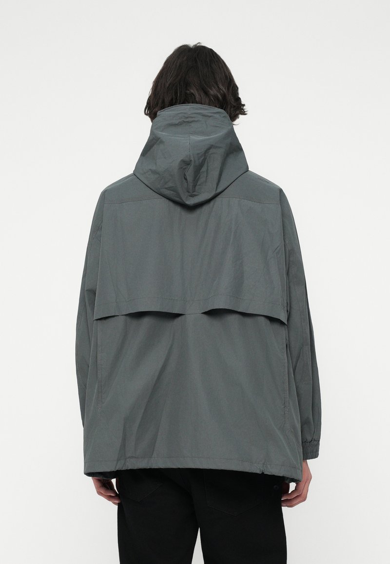 Persoon in een losse, donkergrijze hoodie gezien vanaf de achterkant tegen een effen lichte achtergrond.