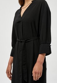 Zwarte jurk met een V-halslijn, lange mouwen die bij de manchetten zijn gathered, en een gladde, gestructureerde stof. Heeft een centrale knoopdetail.