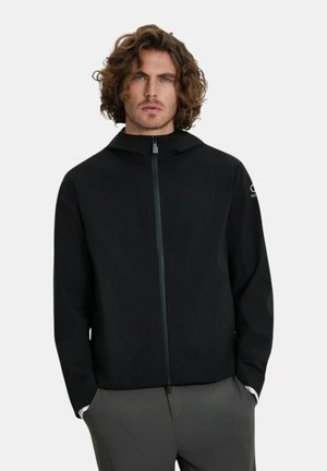 Uomo con capelli ricci che indossa una giacca nera con cappuccio e zip e pantaloni grigi, mani nelle tasche, sfondo neutro.