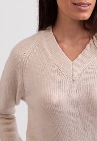 Beige gestrickter Pullover mit V-Ausschnitt, der über einen gerippten Kragen und eine dezente Textur verfügt. Nahaufnahme, die die Details des Stoffes hervorhebt.
