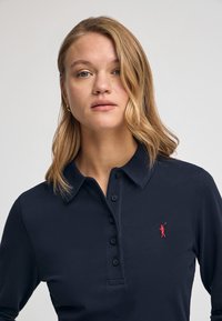 Navyblauer Langarm-Poloshirt mit einem Kragen, einer Dreiknopf-Leiste und einem roten gestickten Logo auf der linken Brust. Glattes, texturiertes Gewebe.