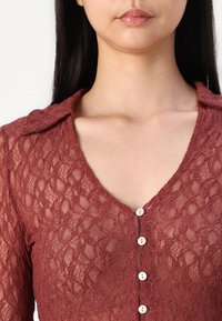Burgundská krajková blůza s výstřihem do V, s límečkem a pěti bílými knoflíky vpředu. Složitý texturovaný vzor po celém povrchu.