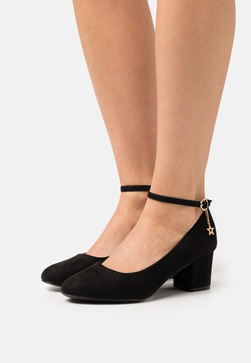 Zapatos de ante negro con tacón bloque y tira en el tobillo que presenta un charm de estrella. Punta redonda y textura suave. Diseño simple con acentos mínimos.