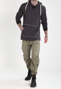 Zwarte hoodie met een voorzak en contrasterende capuchon, gecombineerd met loszittende olijfgroene cargobroeken met meerdere zakken en ritsen. Zwarte laarzen.