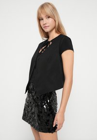 Joven mujer con cabello rubio que lleva una camiseta negra de manga corta, un chaleco negro abotonado y una minifalda negra con lentejuelas, de pie contra un fondo blanco.