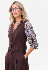 Brązowa kamizelka w paski nad z wzorem kwiatowym bluzki z niebieskimi i różowymi akcentami. Modelka nosi oversize'owe okulary i złote kolczyki w kształcie obręczy.