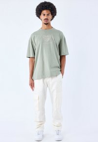 Lichtgroene oversized katoenen t-shirt met een hartgrafiek en "PROJECT X PARIS" tekst, gecombineerd met crèmekleurige cargobroek en witte sneakers.