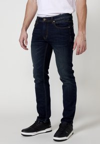 Jeans in denim blu scuro con vestibilità slim, dotati di cinque tasche, chiusura anteriore con bottone e cuciture a contrasto gialle. Indossati con sneaker nere.