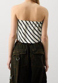 Haut sans manches de couleur crème avec des rayures noires diagonales, associé à un pantalon cargo vert foncé avec plusieurs poches et des détails de couture visibles.