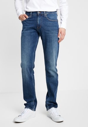Personne portant un jean bleu moyen à jambes droites, des baskets blanches et une chemise blanche à manches longues, debout avec une main dans la poche.
